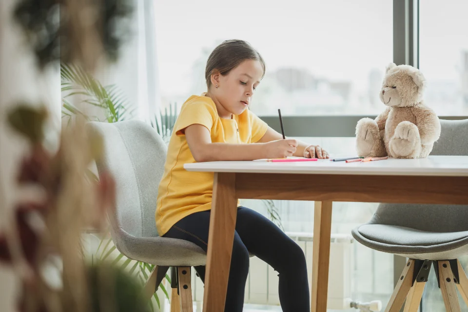 Child writing at a table beside a stuffed toy.